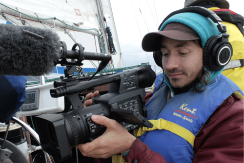 Jamie LeJeune holding a video camera on a boat, wearing headphones and a warm hat and a life vest. 